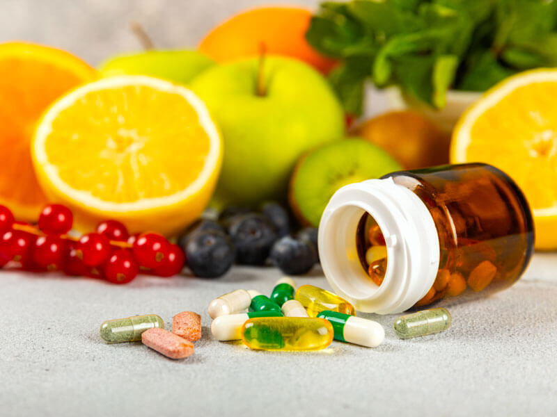 Variety of supplement tablets spilling out of pill bottle in front of fresh fruits and veggies